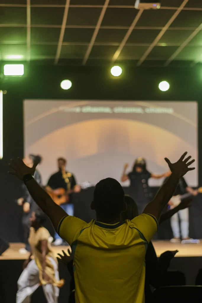Homem canta ao microfone sentado na borda do palco com músicos ao fundo.