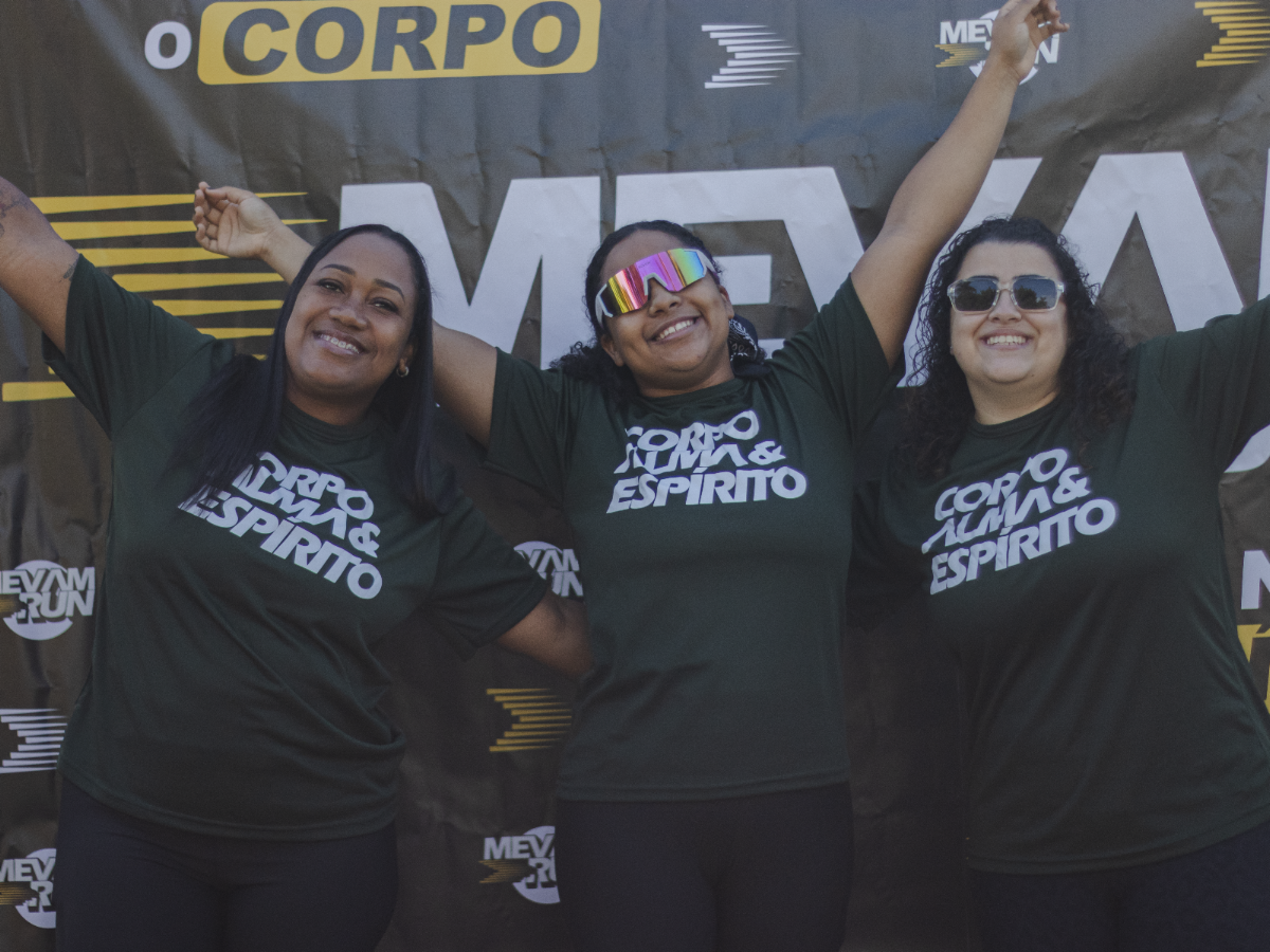 Três participantes do MEVAM RUN com camisetas verdes diante do painel do evento.